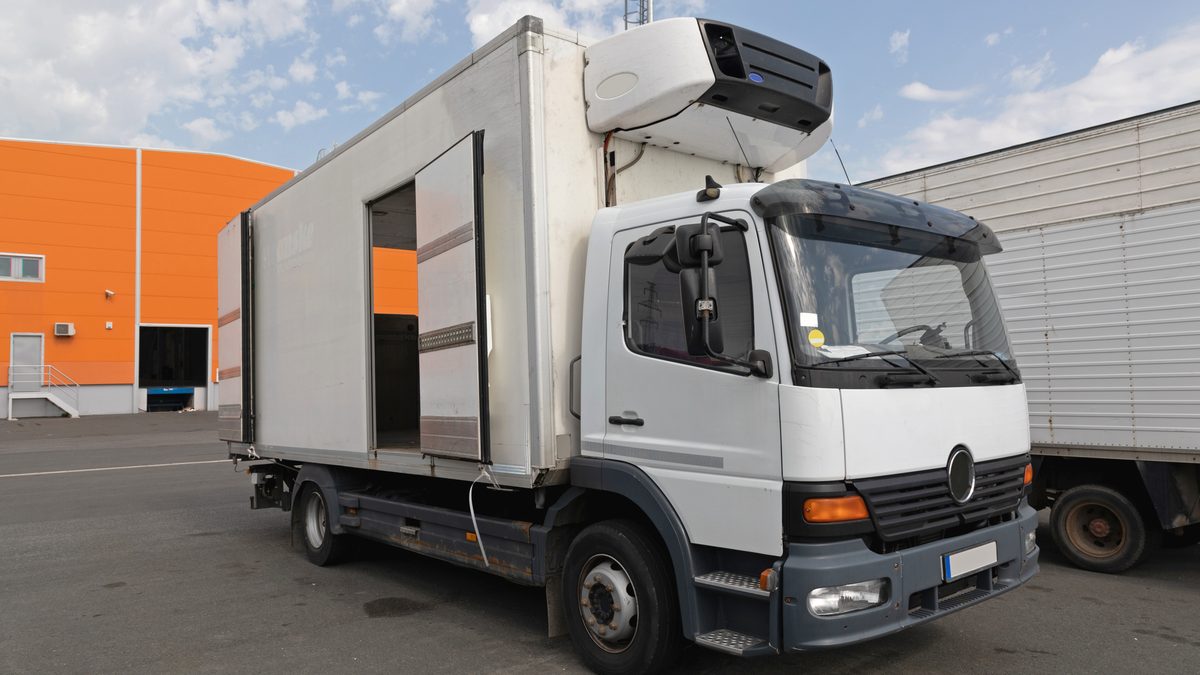 A white refrigerated truck parked in an industrial yard, its refrigeration unit mounted on the front of the cargo box above the cab.