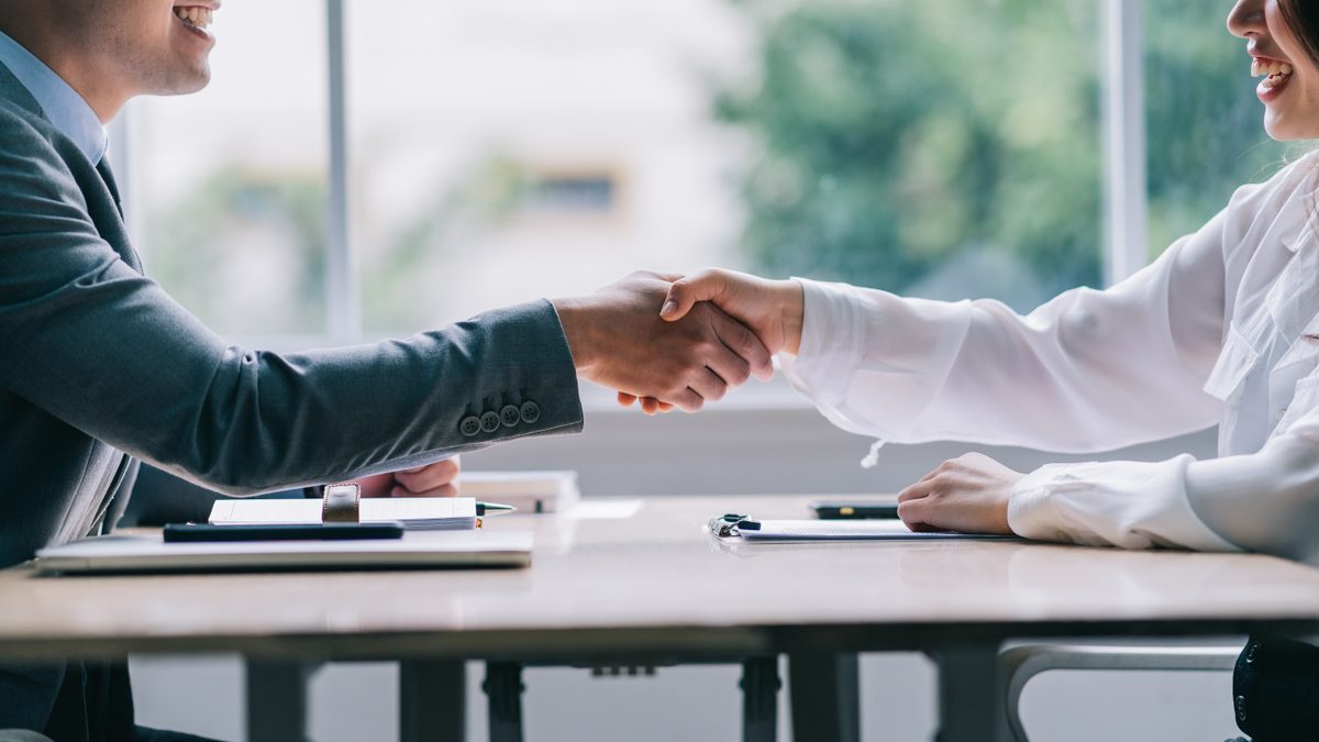 A businessman in a grey suit and a businesswoman in a white blouse shake hands across a desk in a sunlit office.