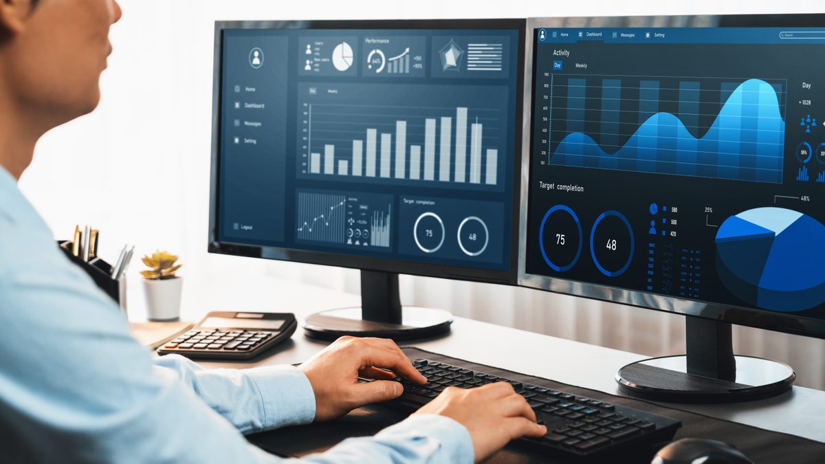 A finance professional works at a desk beneath two large monitors filling the frame with blue business-intelligence dashboards of bar charts, trend lines, and gauge dials.